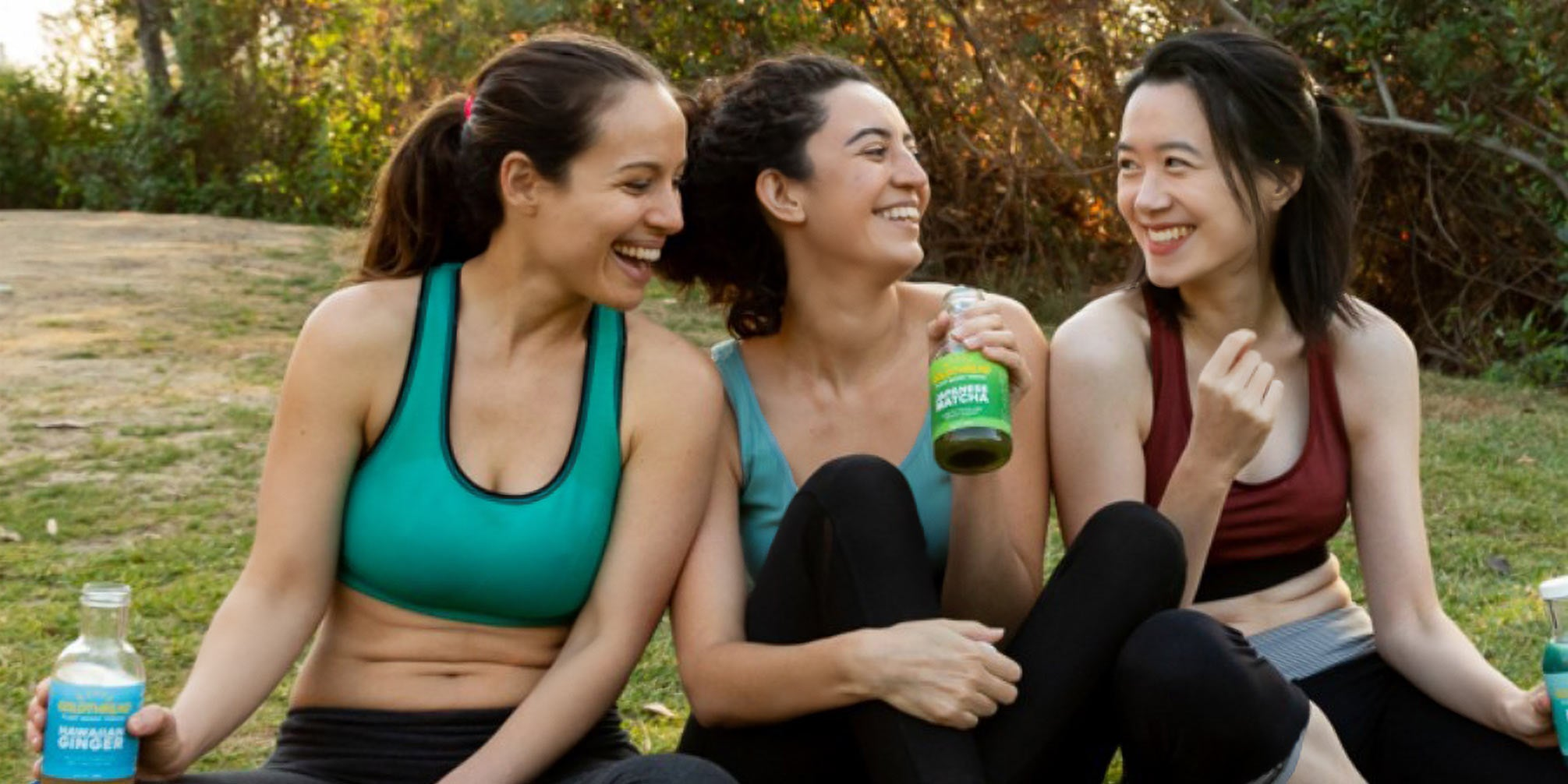 Two women dressed in athletic wear jog side by side, smiling radiantly in a sunlit outdoor setting, fueled by Goldthread tonics.