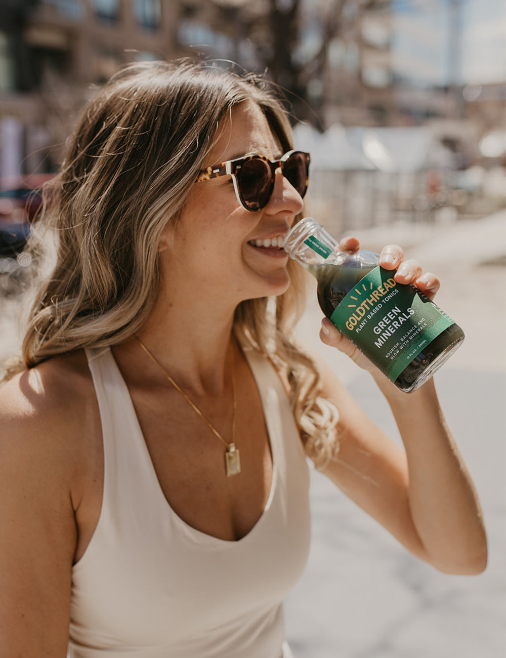 Blond woman wearing sunglasses and drinking Goldthread Green Minerals tonic outside on a sunny day
