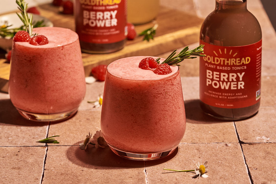 Goldthread Plant-Based Tonics Frozen Boozy Berry Lemonade garnished with raspberries and rosemary next to Goldthread Plant-Based Tonics Berry Power tonic bottles