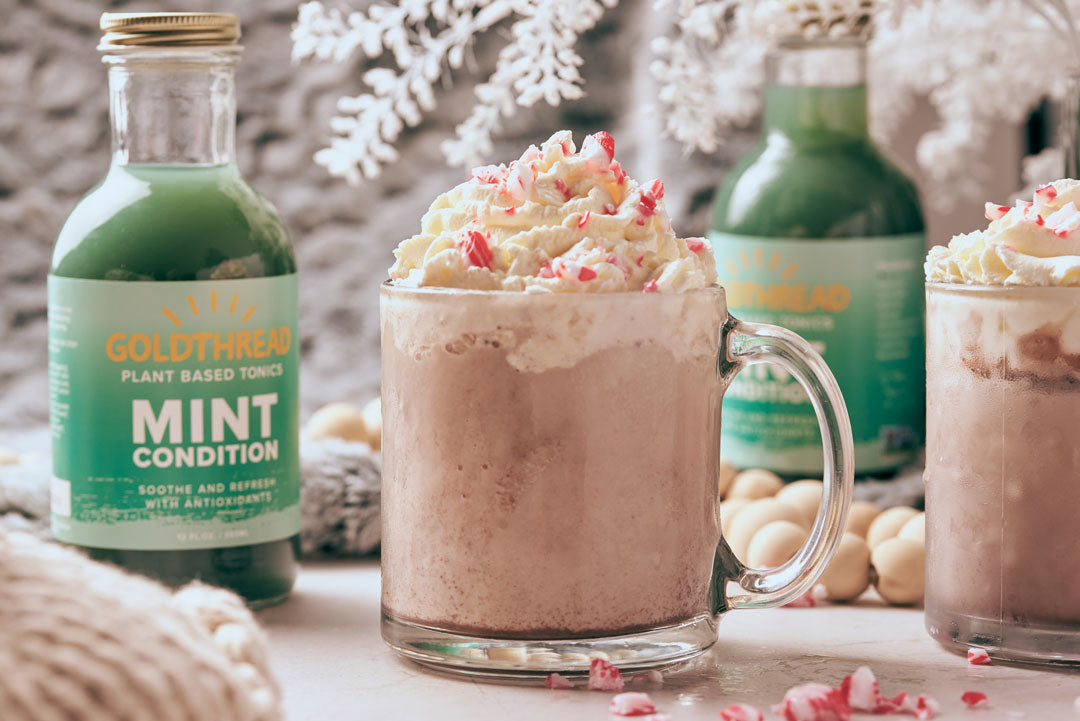 Goldthread Plant-Based Tonics Frozen Mint Hot Chocolate garnished with crushed candy canes and whipped cream with Goldthread Plant-Based Tonics Mint Condition tonic bottles