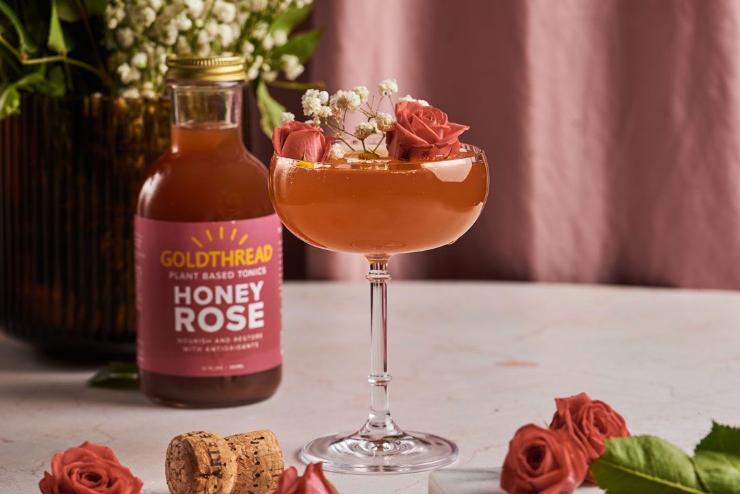 Goldthread Plant-Based Tonics Honey Rose Spritz Cocktail in a coupe glass garnished with roses and chamomile blossoms