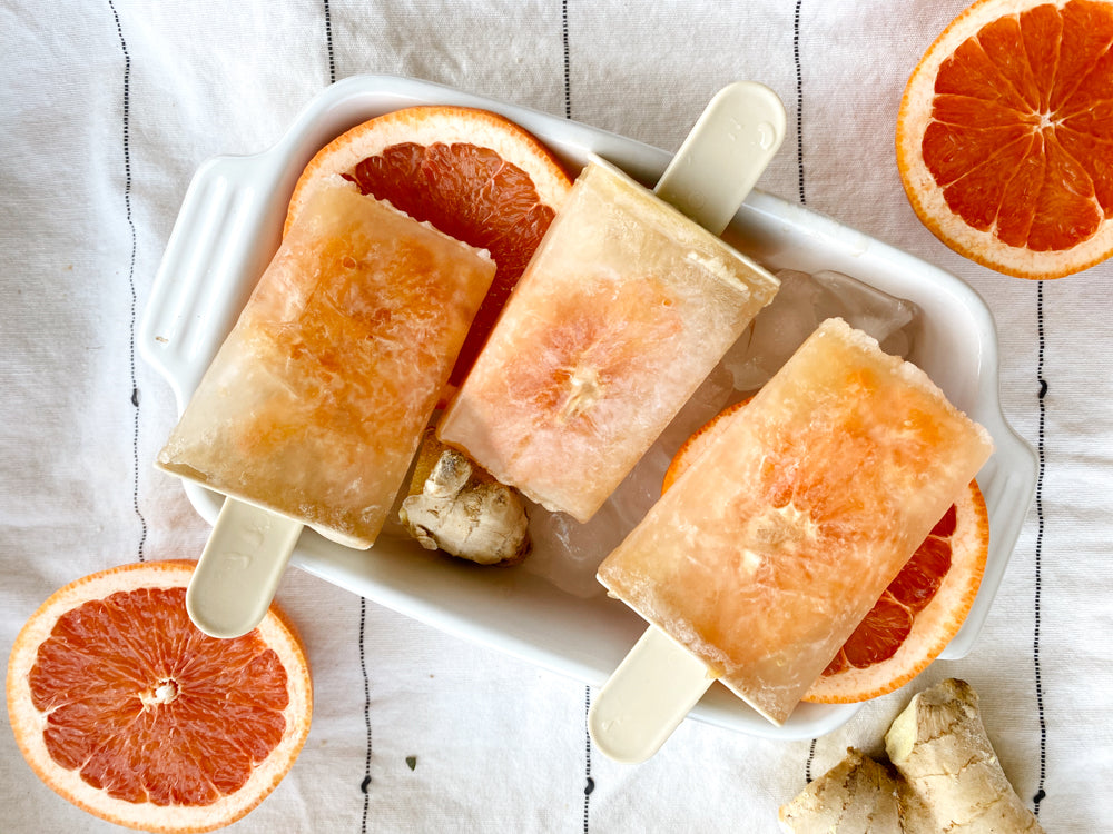 Hawaiian Ginger and Grapefruit popsicles sitting in a white dish on a white tablecloth with slices of grapefruit and ginger pieces