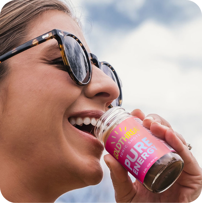 Brunette woman wearing sunglasses and drinking Goldthread Tonics Pure Energy Tonic Shot in an outdoor setting with blue sky in the background