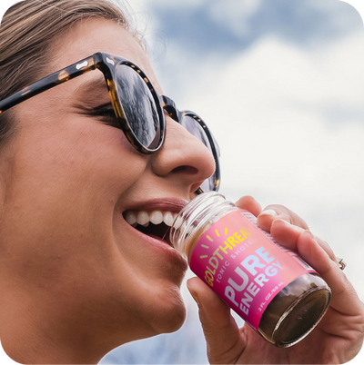Brunette woman wearing sunglasses and drinking Goldthread Tonics Pure Energy Tonic Shot in an outdoor setting with blue sky in the background