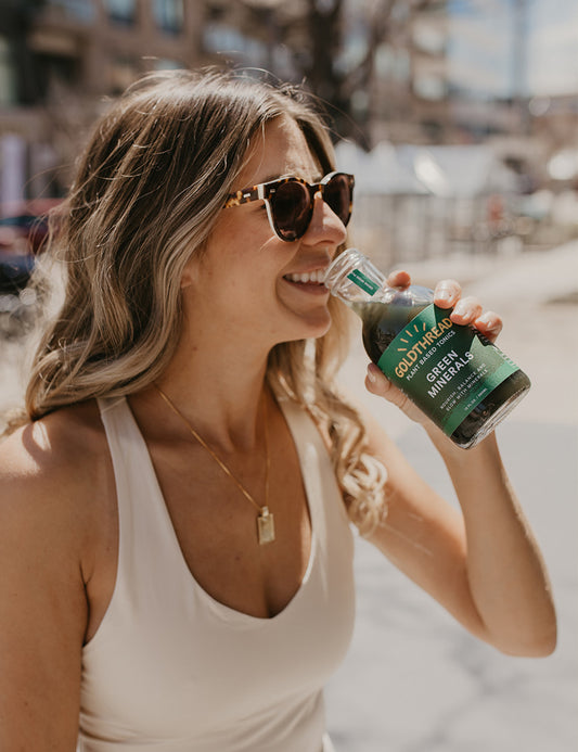 Blond woman wearing sunglasses and drinking Goldthread Green Minerals tonic outside on a sunny day