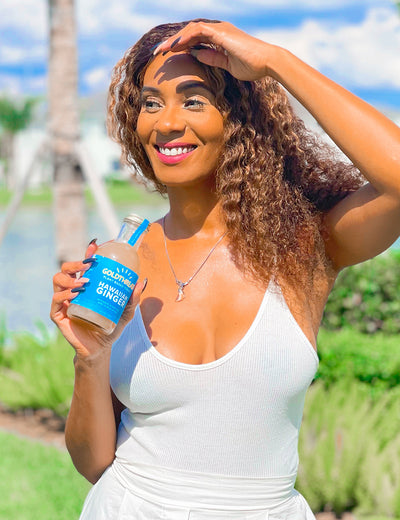 Woman with dark curly hair wearing a white tank top and drinking Goldthread Hawaiian Ginger tonic outside on a sunny day with a palm tree in the background