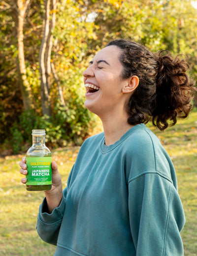 Woman with dark curly hair laughing and holding a bottle of Goldthread Japanese Matcha tonic outside in front of a stand of trees