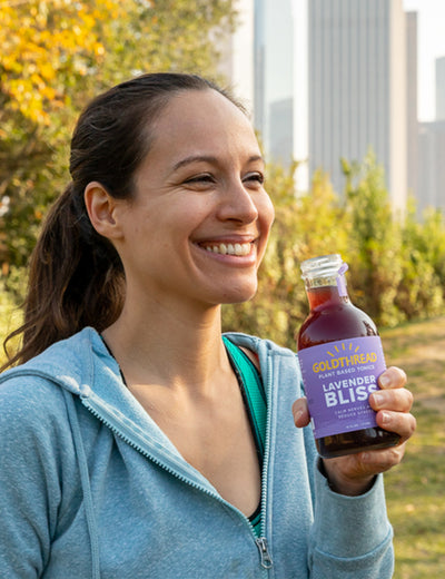 Woman with her hair in a ponytail smiling and holding a bottle of Goldthread Lavender Bliss tonic